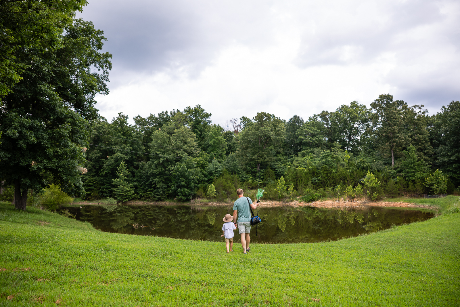 Father and young child holding hands while walking toward a pond with fishing gear on a summer day