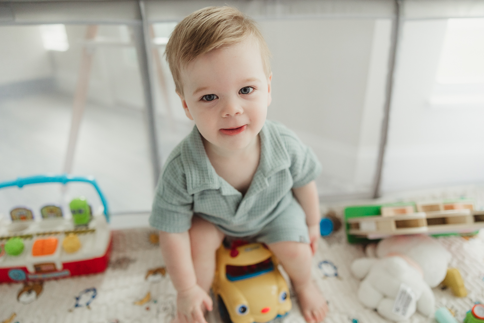 Little boy sitting on a toy truck inside his playpen, surrounded by colorful toys, smiling at the camera during a real life documentary photoshoot.