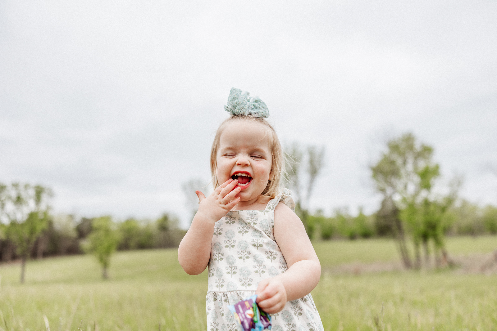 Toddler girl smiling while eating fruit snacks during an outdoor family photography session in Bonne Terre, Missouri.